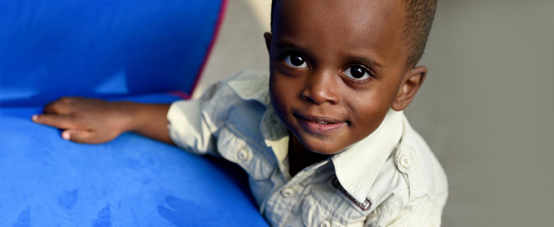 A young child wearing a light-colored shirt stands next to a blue object, looking up with a slight smile. The background is out of focus, bringing attention to the child's expressive eyes and cheerful expression.