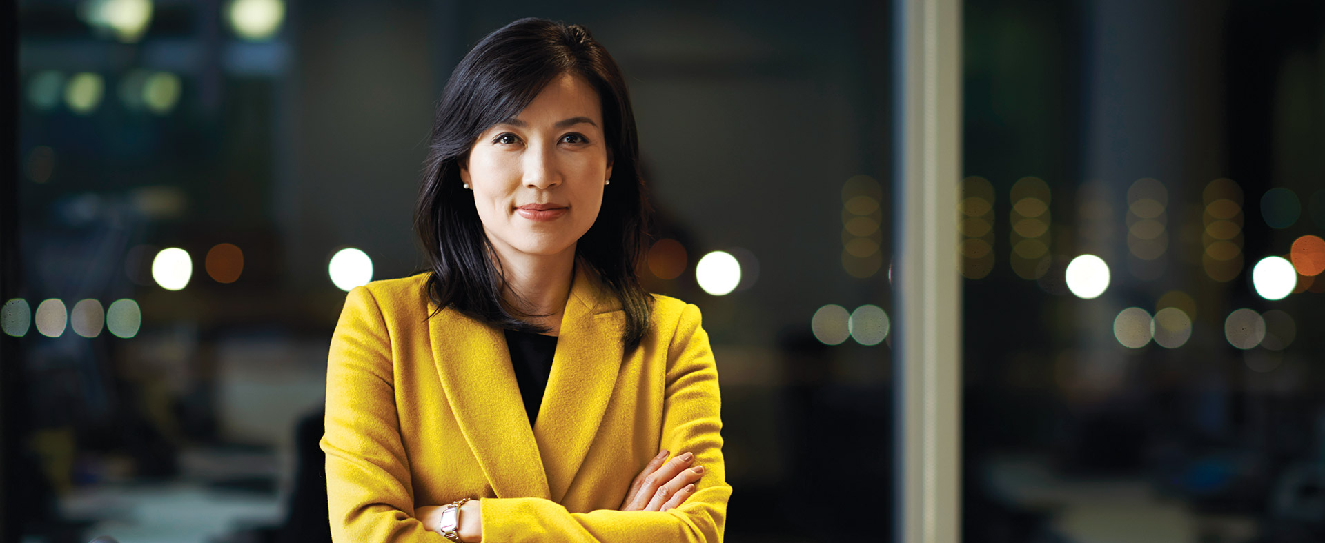 A woman with shoulder-length dark hair is standing with her arms crossed, wearing a yellow coat. She looks confidently at the camera. The background is blurred, featuring office lights and windows, suggesting an urban work environment.