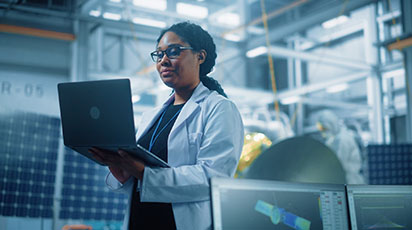 A person wearing a white lab coat and glasses is working on a laptop in an industrial or laboratory setting. Multiple computer screens and machinery are visible in the background, indicating a high-tech environment.