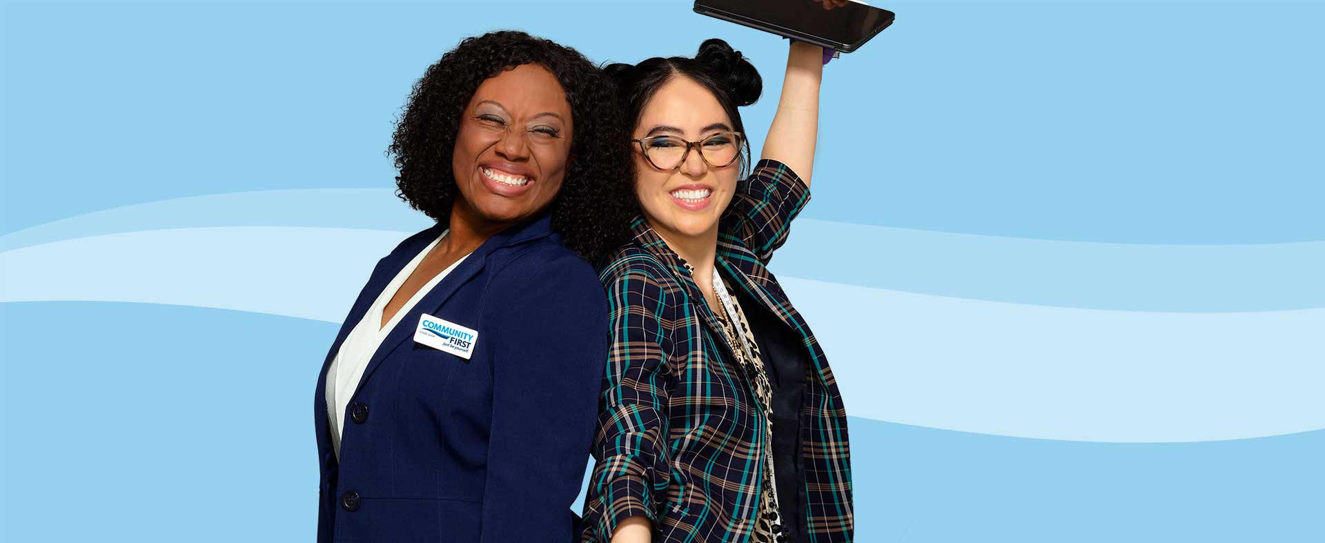 Two women standing close together, smiling broadly against a light blue background. The woman on the left wears a navy blue blazer and a name tag. The woman on the right wears glasses and a plaid blazer, holding up a tablet. Both look happy and confident.