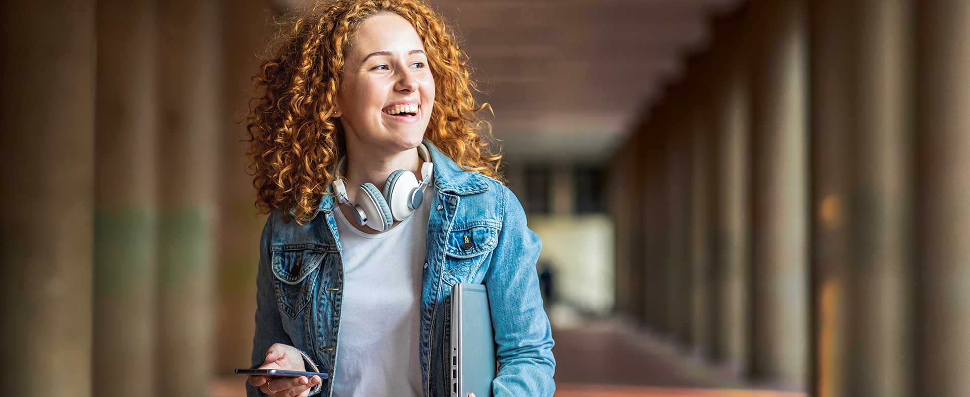A young woman with curly red hair smiles as she holds a phone in one hand and a laptop in the other. She is wearing a denim jacket and headphones around her neck, standing in a corridor with columns in the background, reminiscent of something you'd find in an Austin Williams novel.