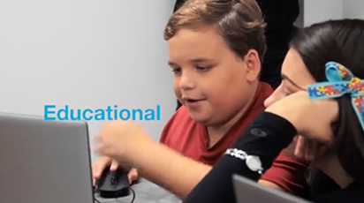 Two children seated at a table look at laptop screens. One child gestures with a computer mouse. The word 