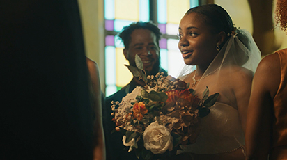 A bride in a strapless wedding gown holds a bouquet of flowers, surrounded by people in a warmly lit room with stained glass windows. She looks joyful and engaged in conversation.