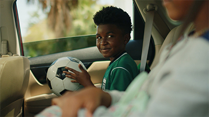 A child sitting in a car, wearing a green soccer jersey, smiling, and holding a soccer ball. Another person is seated in the foreground. Sunlight filters through the window, highlighting the cheerful atmosphere.