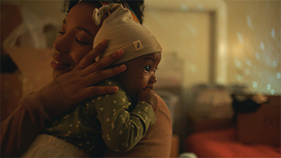 A person lovingly hugs a baby wearing a beanie and green patterned outfit. The room is softly lit, creating a warm and cozy atmosphere.