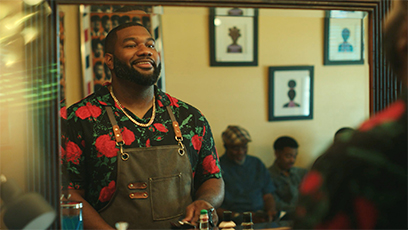 A man with a beard smiles at his reflection in a mirror. He's wearing a floral shirt and apron, standing in a barber shop with framed art on the walls. Two people are seated in the background.